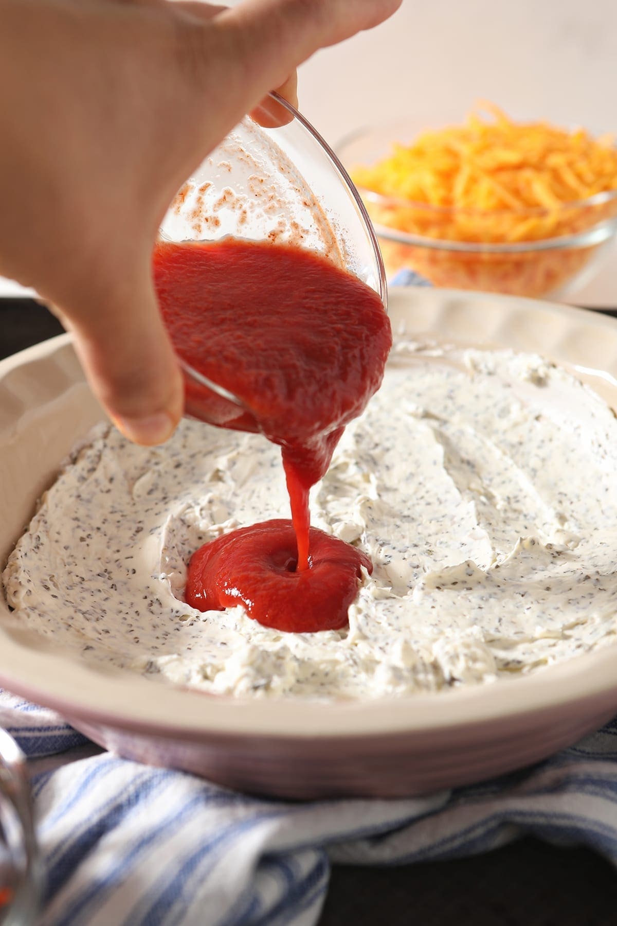 A woman pours pizza sauce on top of a creamy cheese layer in a pie dish