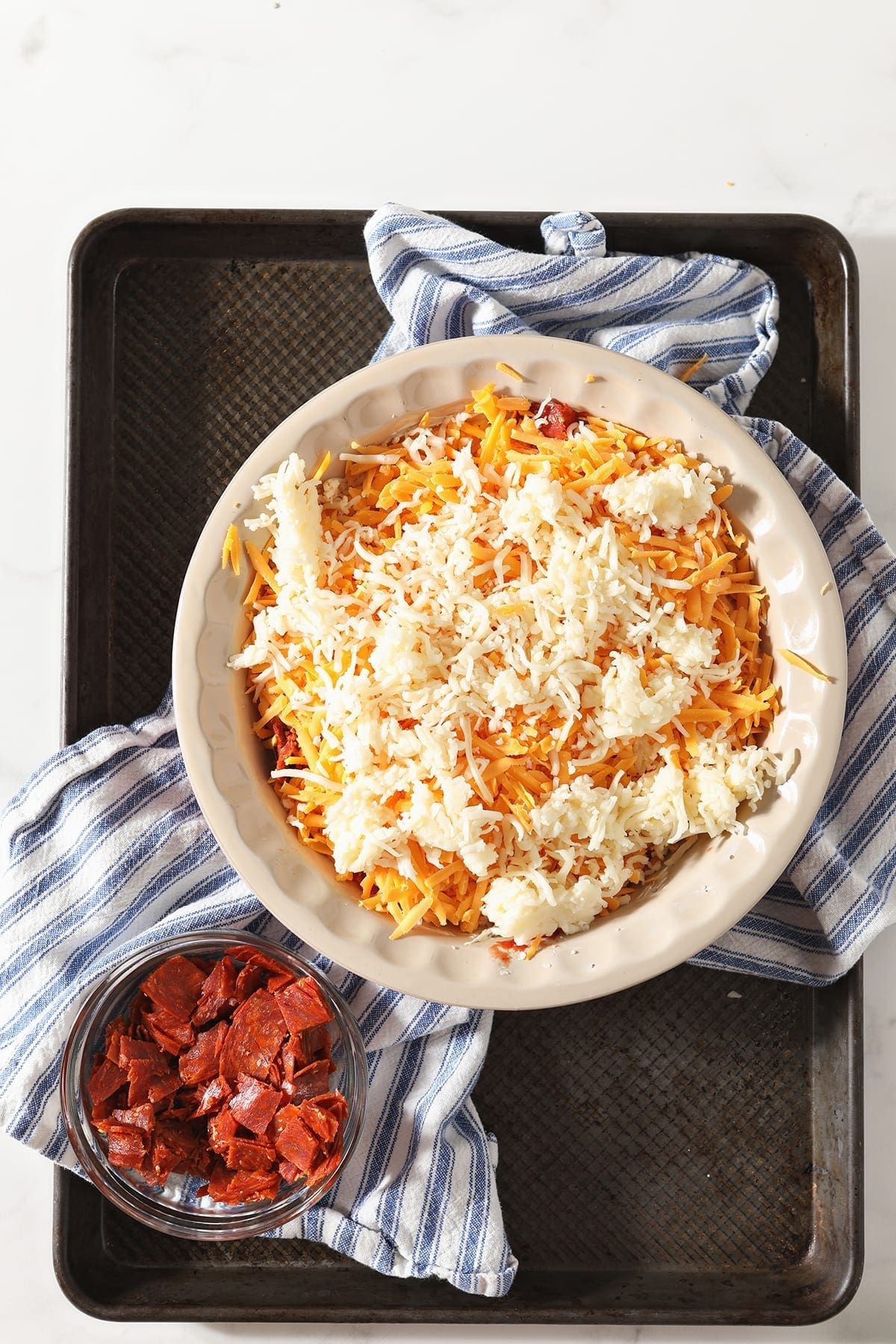 Cheddar and mozzarella cheeses in a pie plate on a blue and white striped towel with a bowl of chopped pepperoni