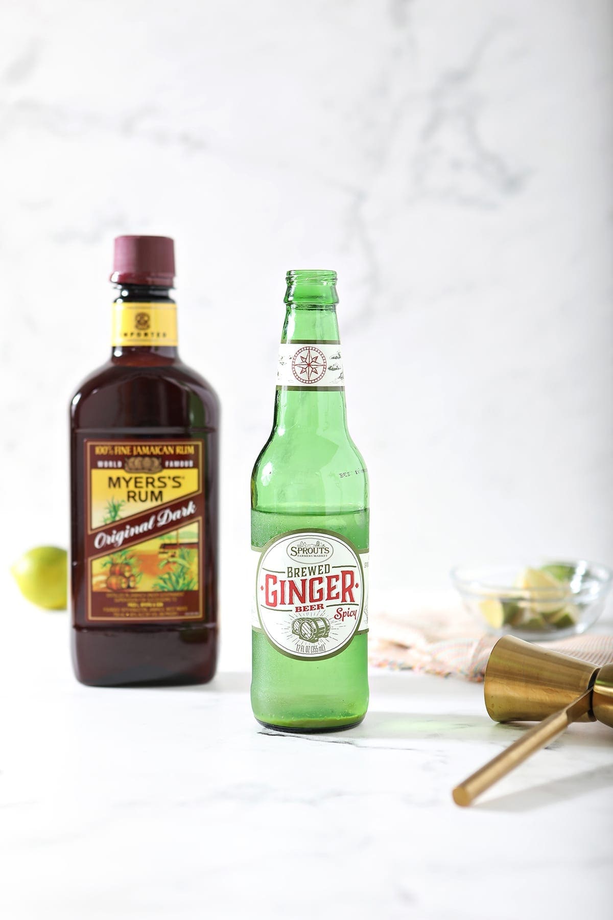 A bottle of ginger beer, a bottle of rum, a jigger and some limes on a marble backdrop