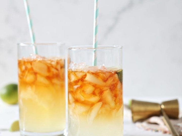Two Dark and Stormy Drinks sit on marble surrounded by ice cubes, lime wedges and a gold jigger