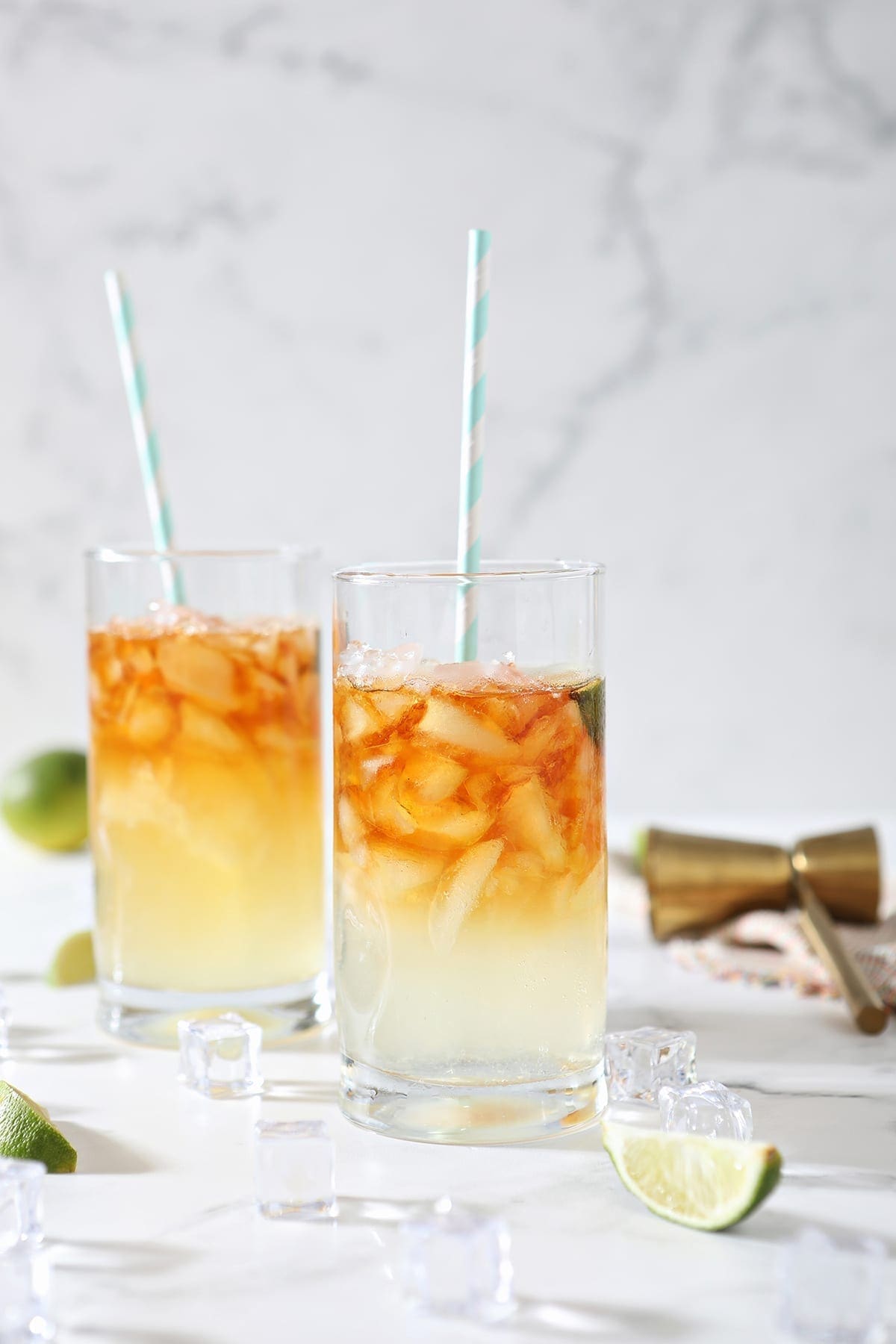 Two Dark and Stormy Drinks sit on marble surrounded by ice cubes, lime wedges and a gold jigger