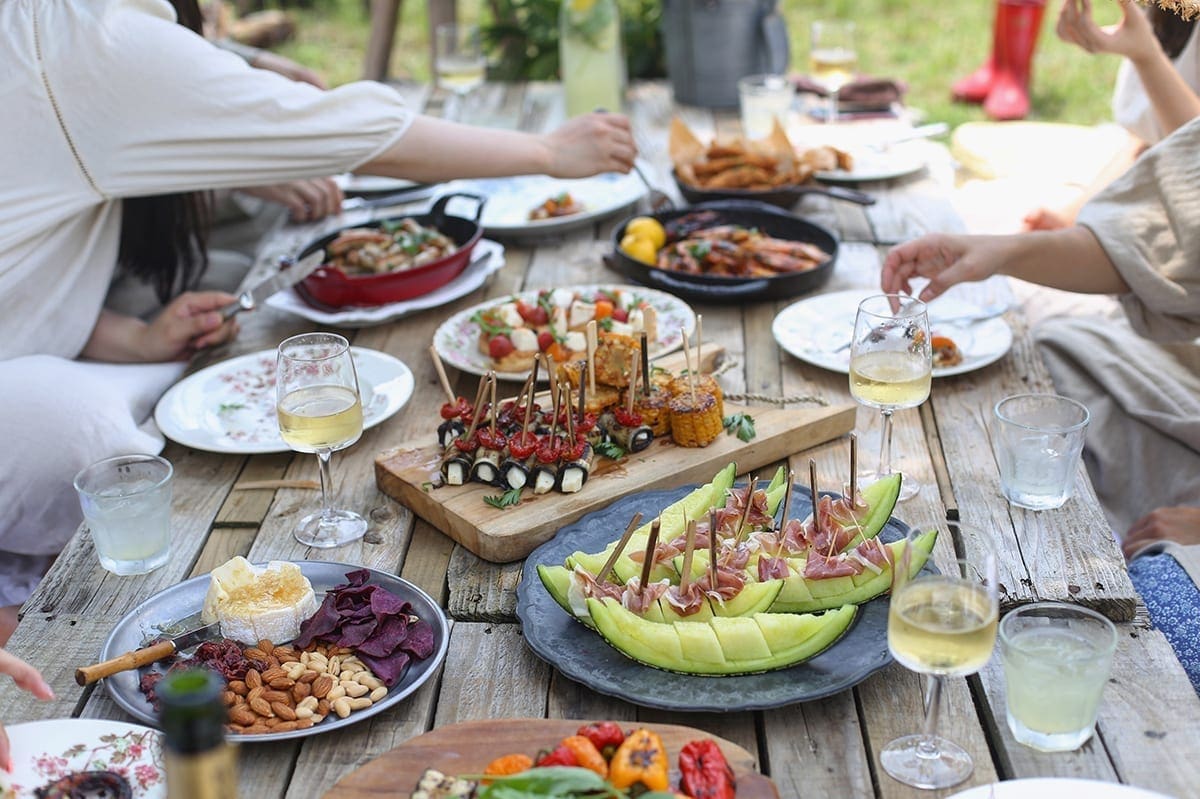 An outdoor table is laden with food and people sit around it