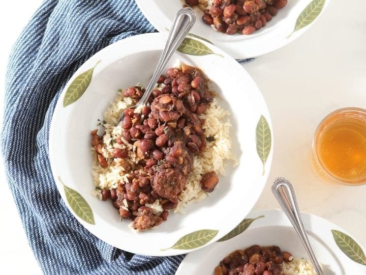 Three decorative white bowls hold servings of Instant Pot Red Beans and Rice, on a blue towel on marble with glasses of beer surrounding