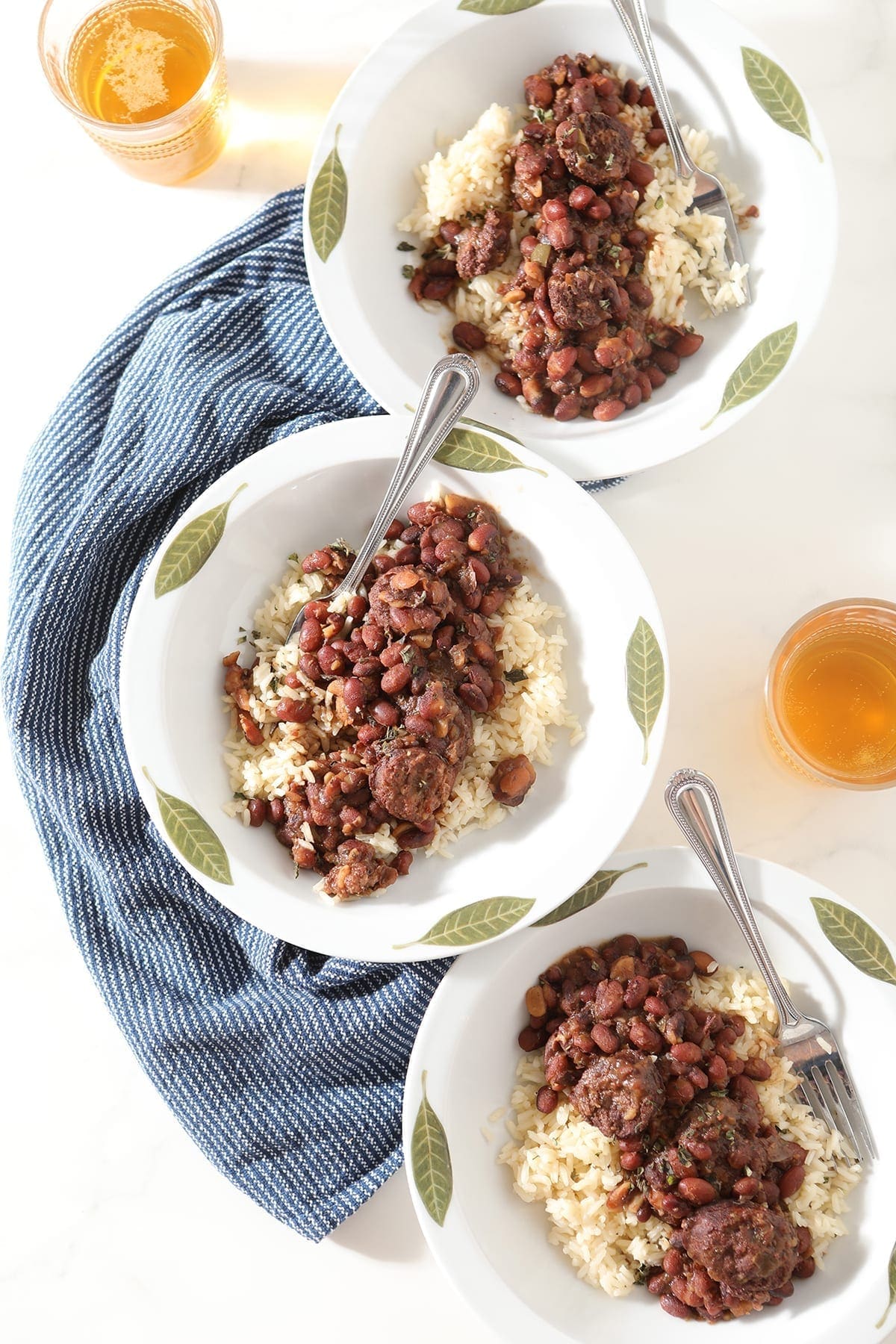 Three decorative white bowls hold servings of Instant Pot Red Beans and Rice, on a blue towel on marble with glasses of beer surrounding