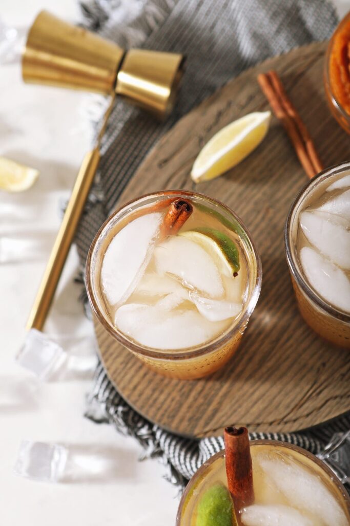 Overhead of three Pumpkin Spice Moscow Mules on a wooden serving dish next to to limes, a jigger and a bowl of pumpkin puree