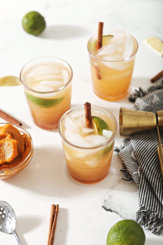 Three Pumpkin Mules, garnished with cinnamon sticks and lime wedges, surrounded by limes, pumpkin and a jigger on marble