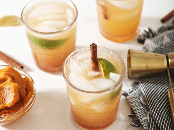 Three Pumpkin Mules, garnished with cinnamon sticks and lime wedges, surrounded by limes, pumpkin and a jigger on marble