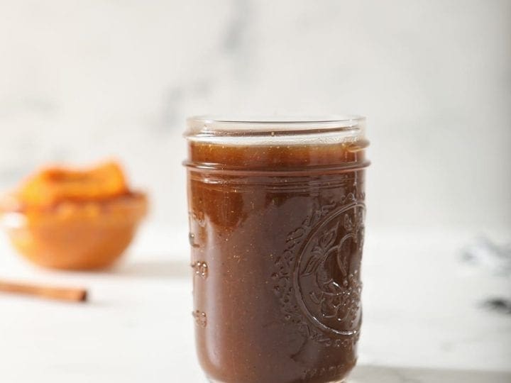 A jar of Homemade Pumpkin Spice Syrup on marble with a bowl of pumpkin puree and a stick of cinnamon behind it