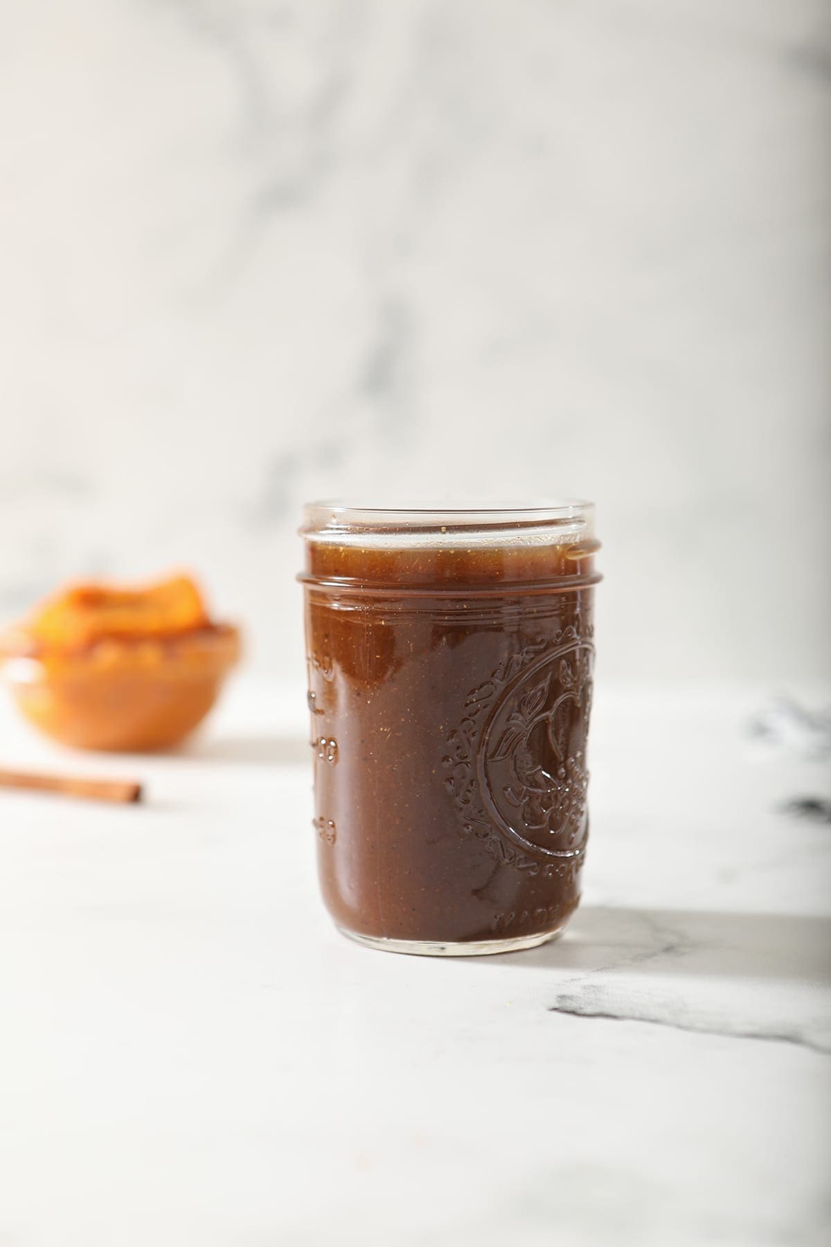 A jar of Homemade Pumpkin Spice Syrup on marble with a bowl of pumpkin puree and a stick of cinnamon behind it