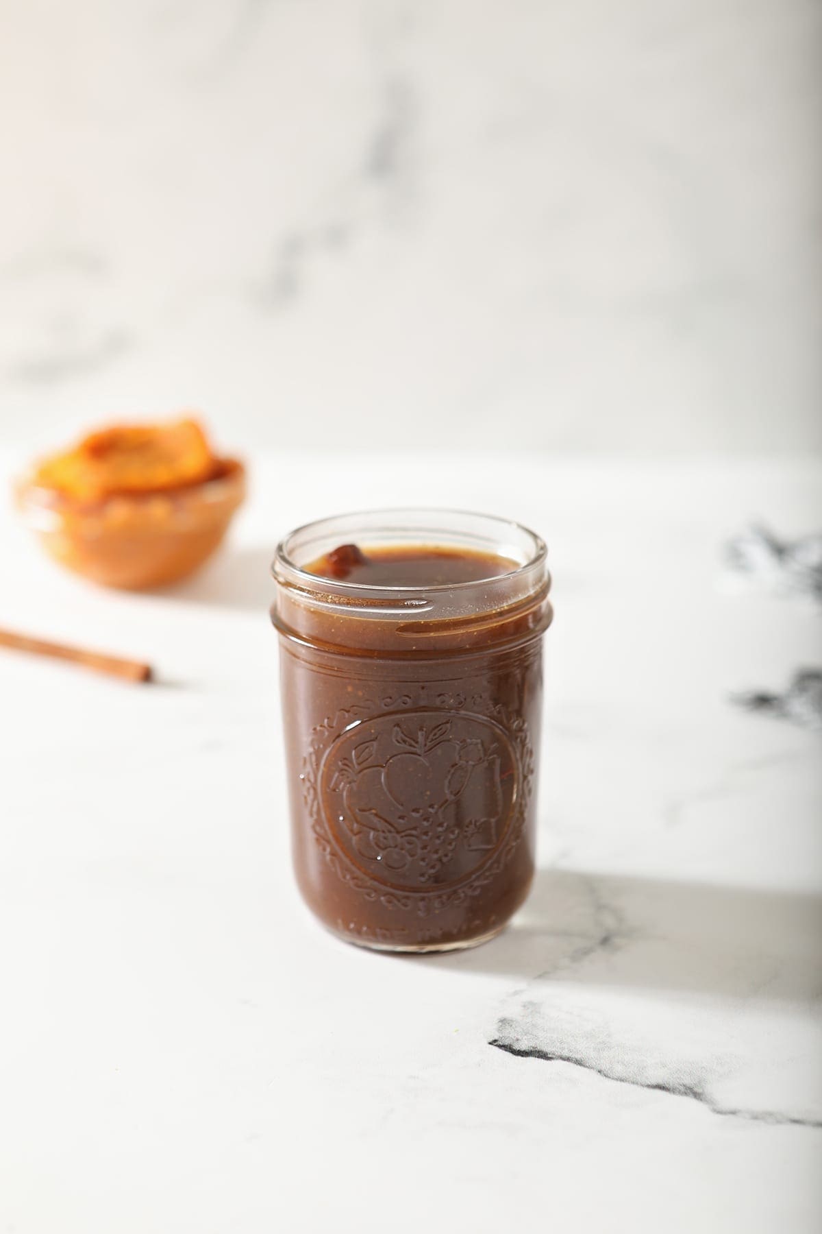 A jar of Pumpkin Spice Syrup sits on marble with a cinnamon stick and pumpkin puree behind it