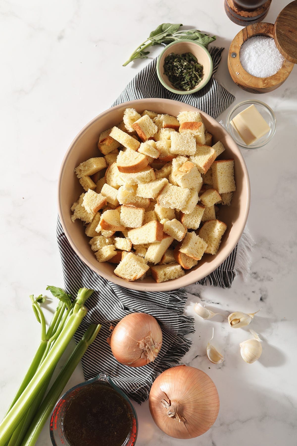 Cubed bread, onions, celery, garlic, vegetable stock and spices on a gray striped towel on marble