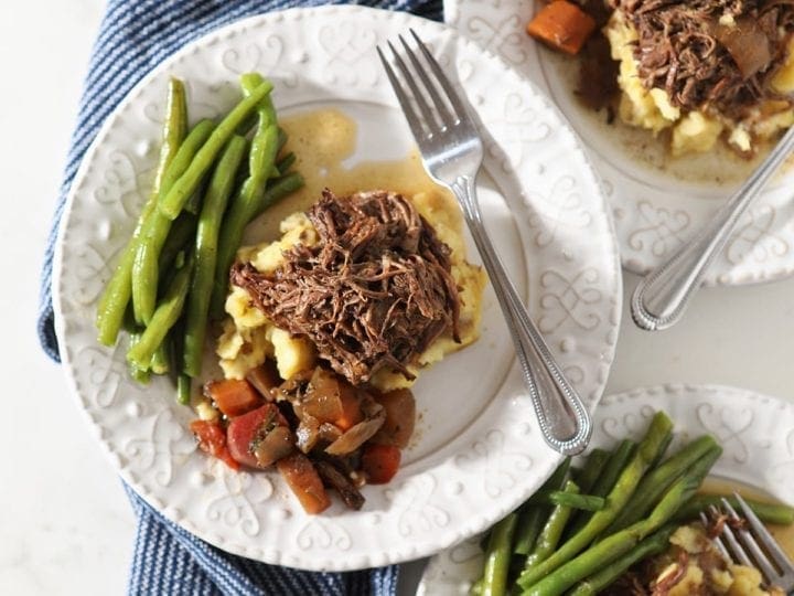 Three plates hold shredded slow cooker pot roast with mashed potatoes, steamed green beans and slow cooked veggies from above