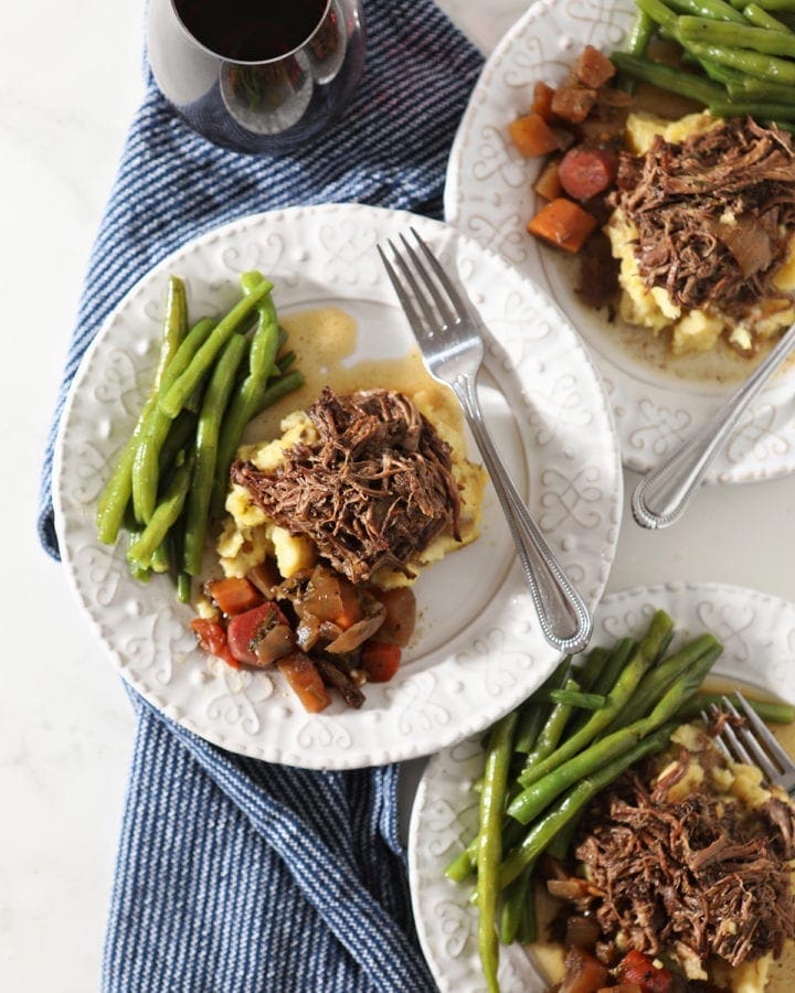 Three plates hold shredded slow cooker pot roast with mashed potatoes, steamed green beans and slow cooked veggies from above
