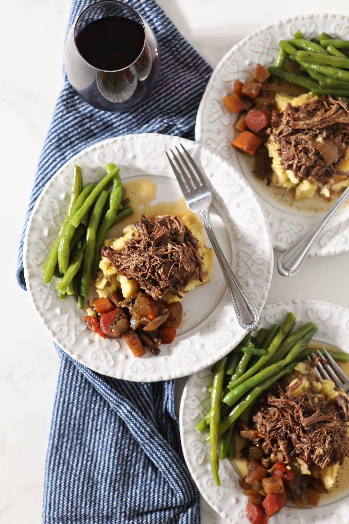 Three plates hold shredded slow cooker pot roast with mashed potatoes, steamed green beans and slow cooked veggies from above