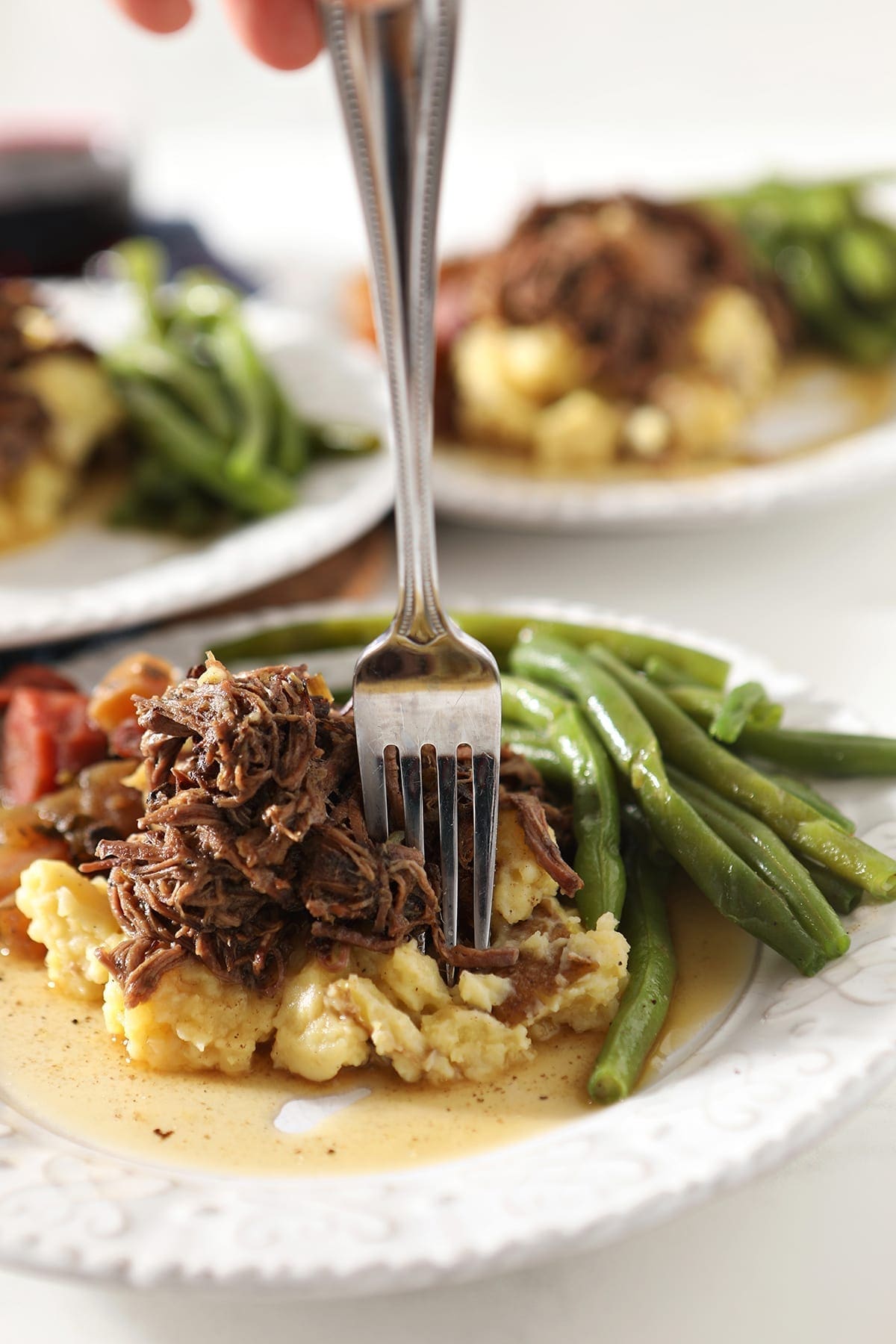 Close up of a fork sticking shredded slow cooker pot roast on top of mashed potatoes and next to steamed green beans