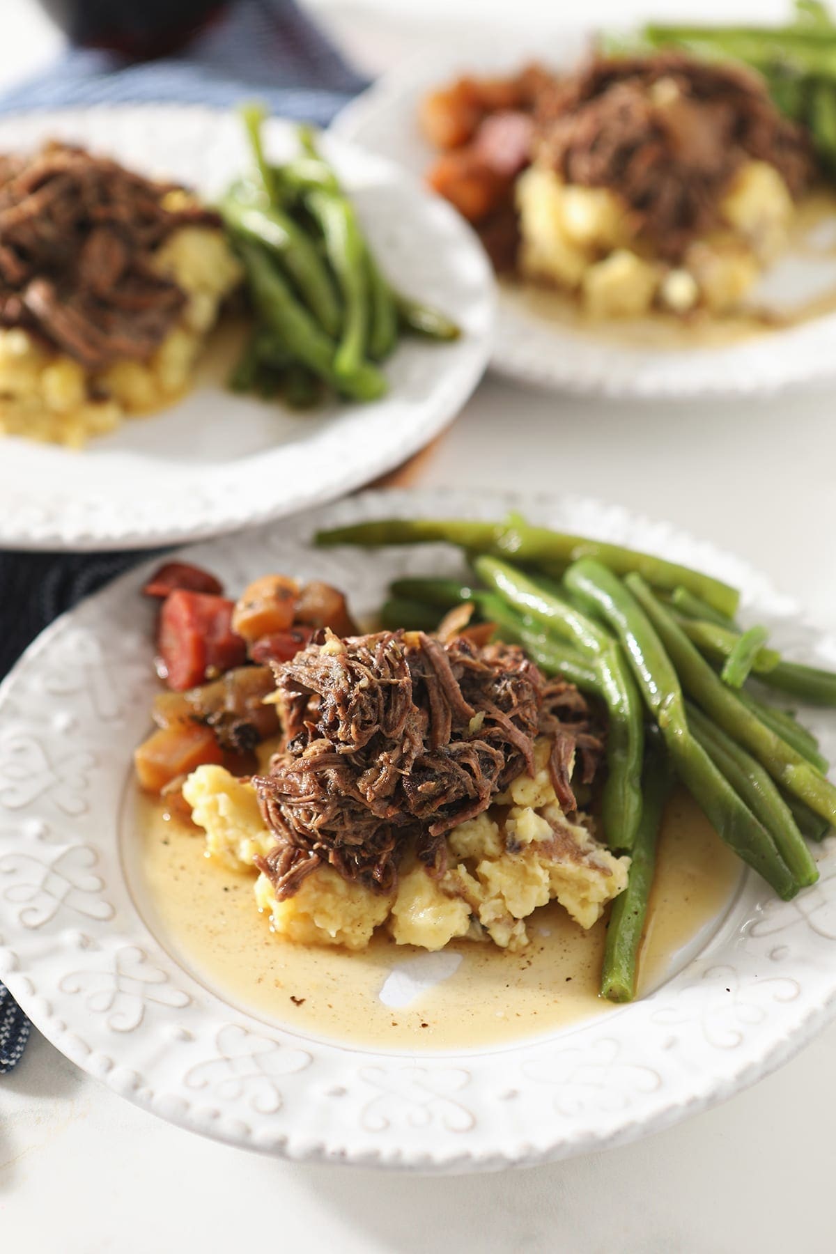 Three plates hold shredded slow cooker pot roast with mashed potatoes, steamed green beans and slow cooked veggies from the side