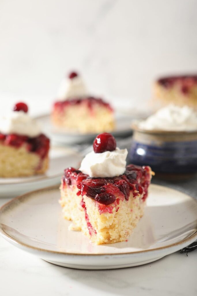 A slice of cranberry upside down cake garnished with a dollop of whipped cream and a cranberry on a white pottery plate with other slices behind it