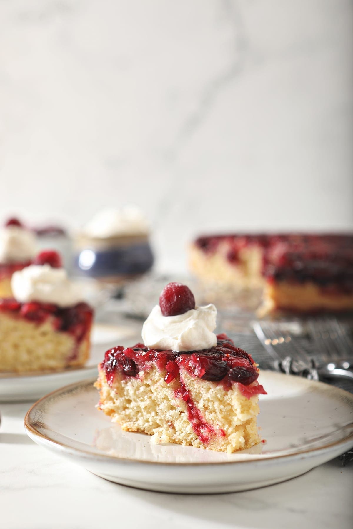 A slice of cranberry upside down cake garnished with a dollop of whipped cream and a cranberry on a white pottery plate with other slices behind it 