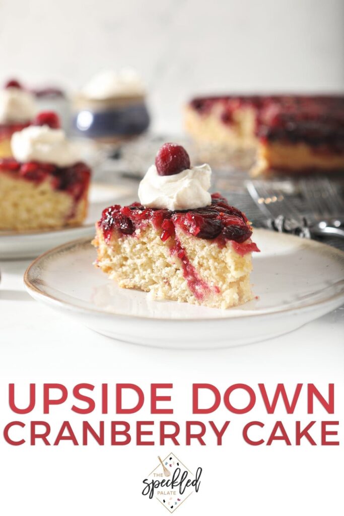 A slice of cranberry upside down cake garnished with a dollop of whipped cream and a cranberry on a white pottery plate with other slices behind it with the text 'upside down cranberry cake'