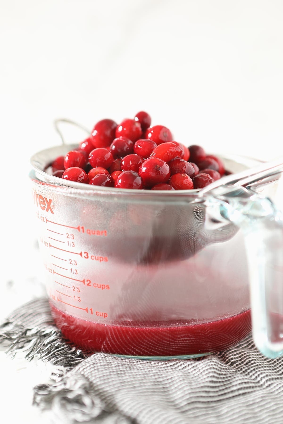 A fine mesh strainer holds cranberries as juices drip into a pyrex glass measuring cup