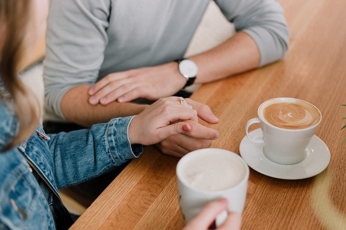 A man and a woman hold hands as they drink coffees at a wooden table