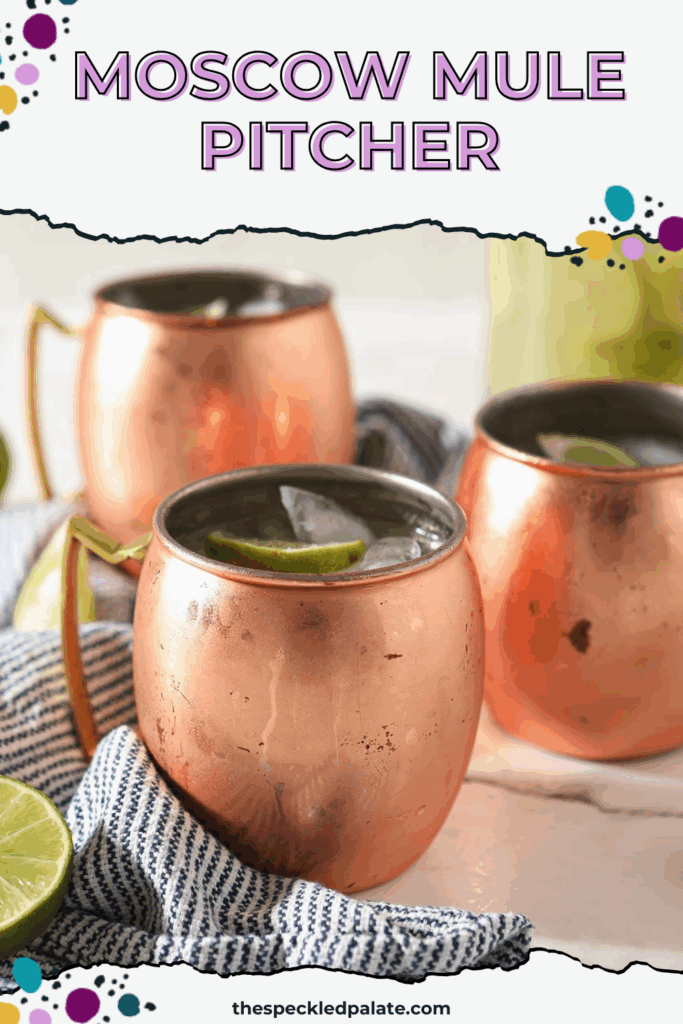 Three copper mugs sitting on a blue and white striped towel hold Moscow mules garnished with lime wedges on marble with a pitcher behind them with the text 'moscow mule pitcher'