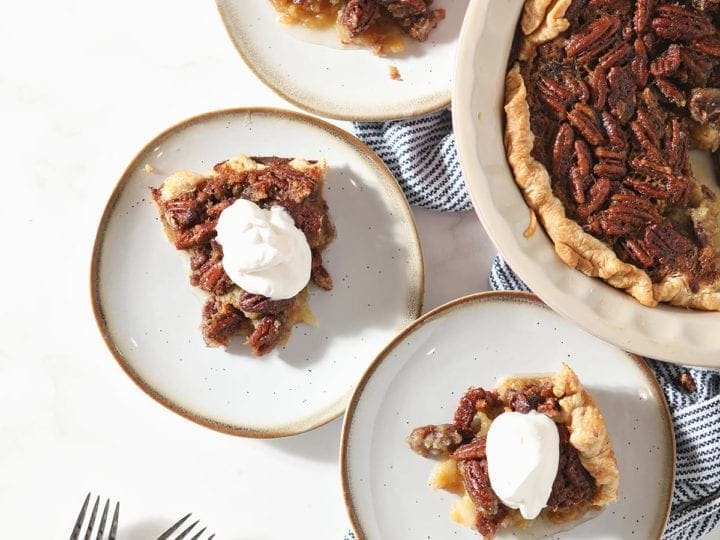 Three slices of Bourbon Pecan Pie sit on white plates next to the rest of the pie, a bowl of whipped cream and a plate holding the serving fork