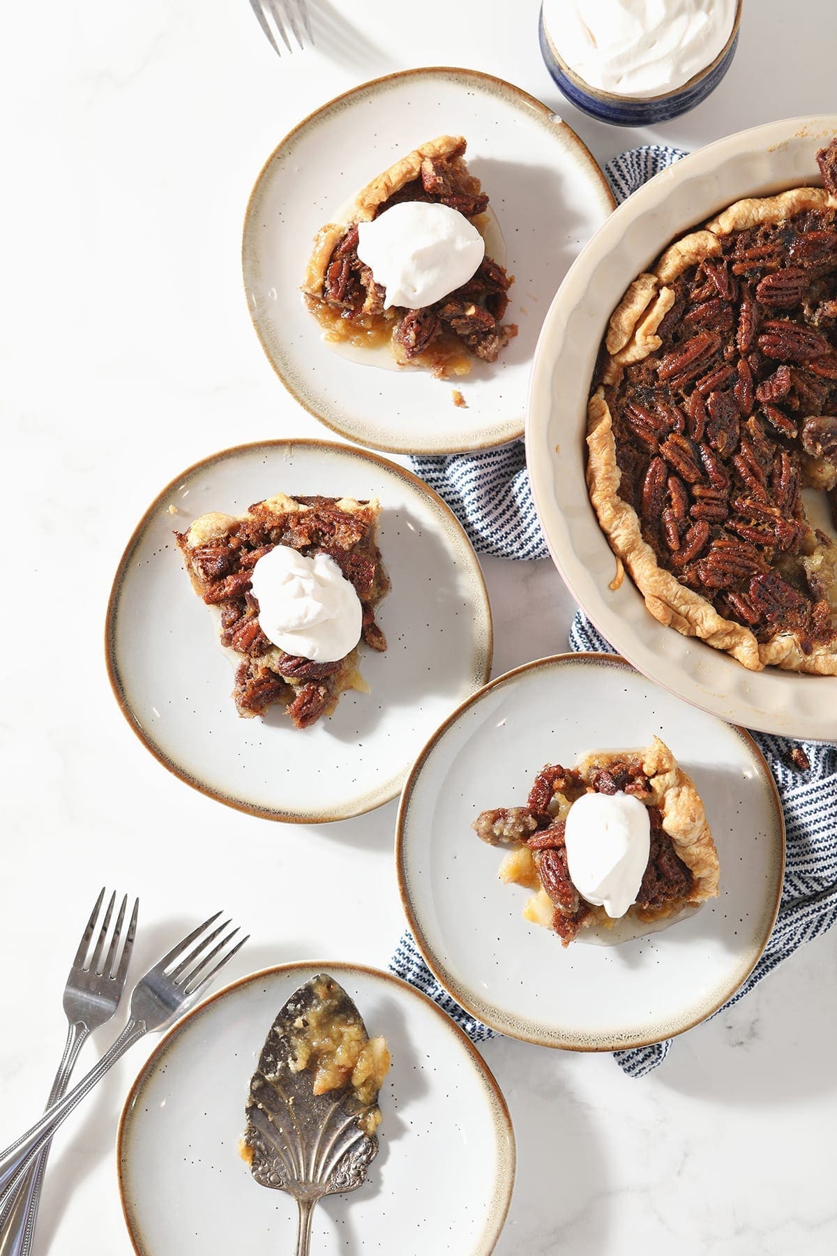 Three slices of Bourbon Pecan Pie sit on white plates next to the rest of the pie, a bowl of whipped cream and a plate holding the serving fork