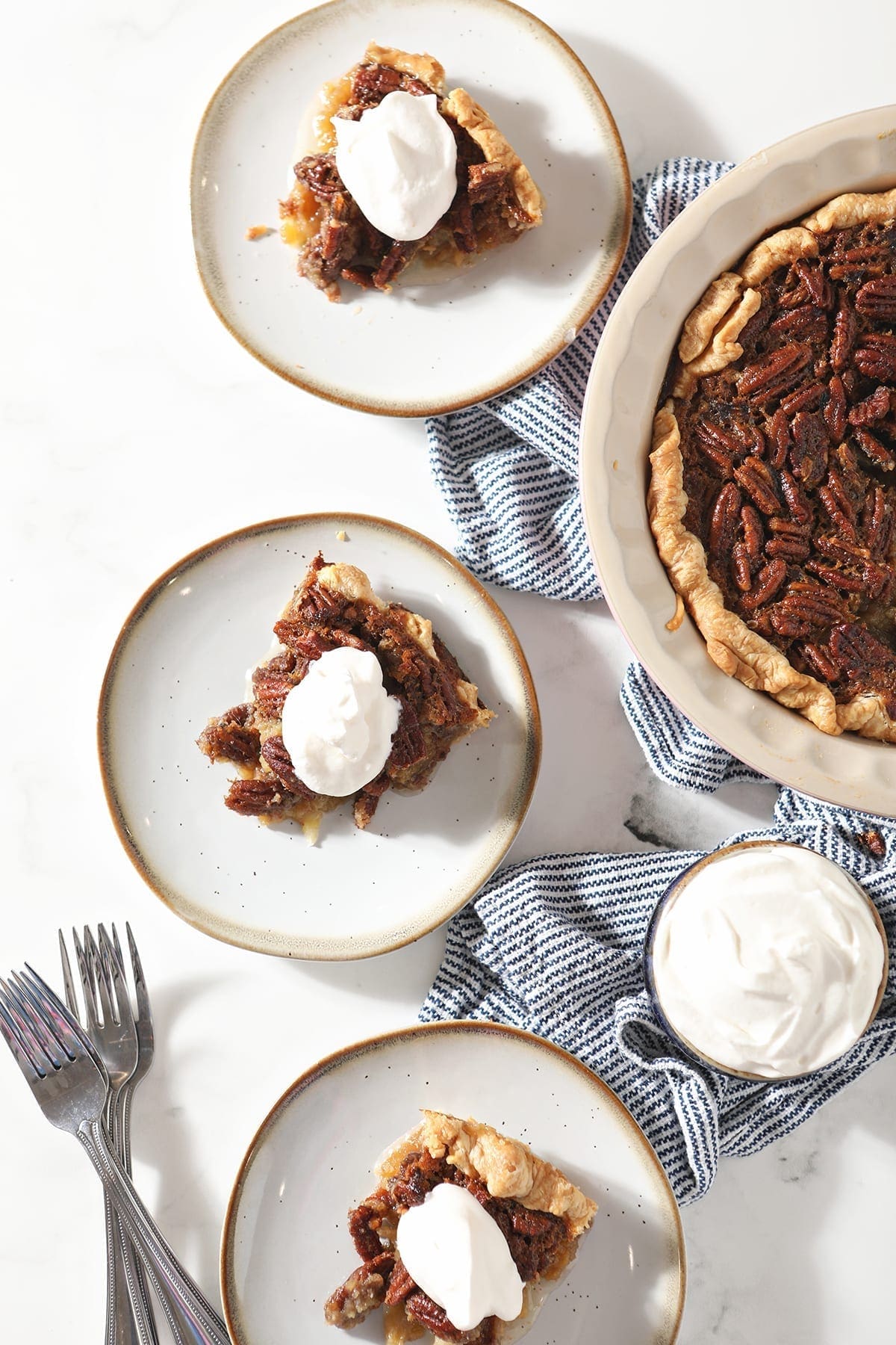 Overhead of three slices of Bourbon Pecan Pie on plates with forks next to the rest of the pie and a bowl of whipped cream