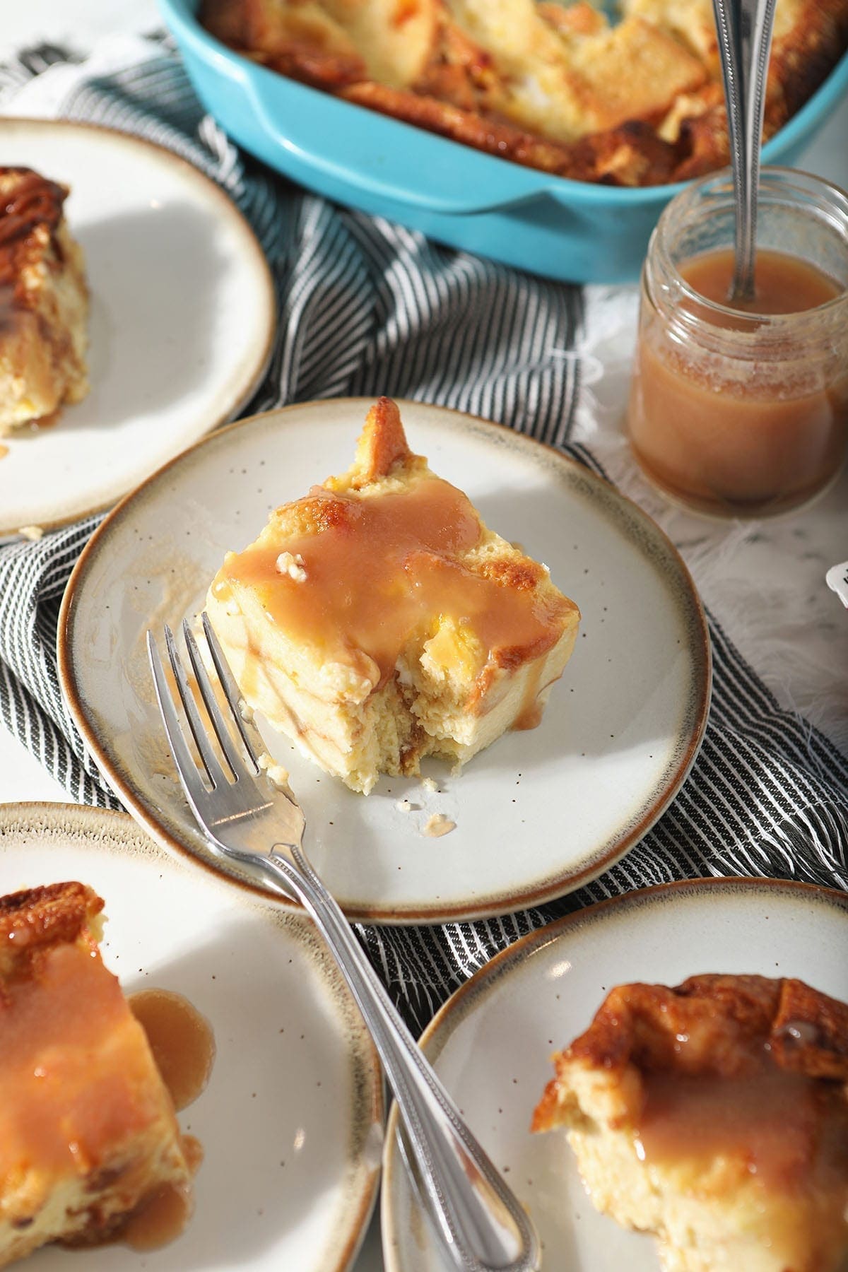 A cut into piece of bread pudding with caramel sauce on a white plate, surrounded by other slices of the dessert and a jar of caramel sauce