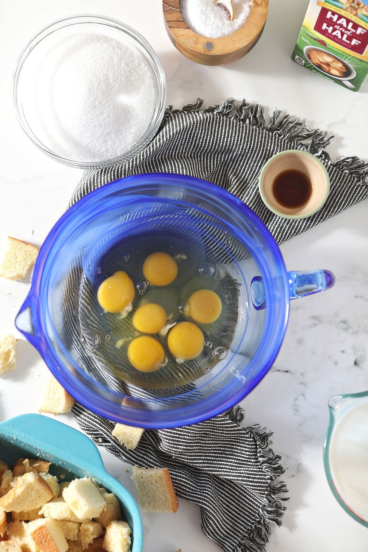 Eggs in a blue bowl surrounded by sliced bread, sugar, vanilla, salt and half and half