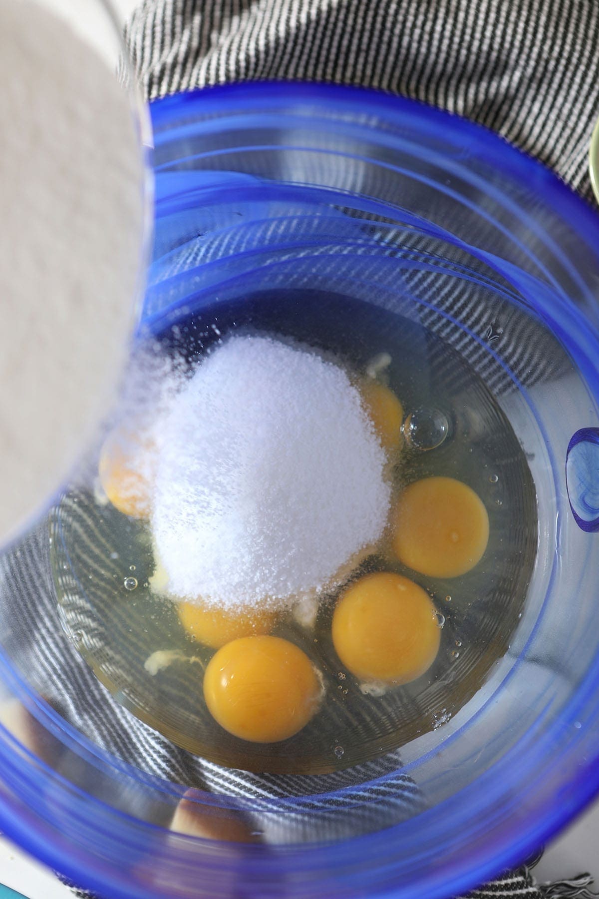 Sugar is poured on top of eggs in a blue glass bowl