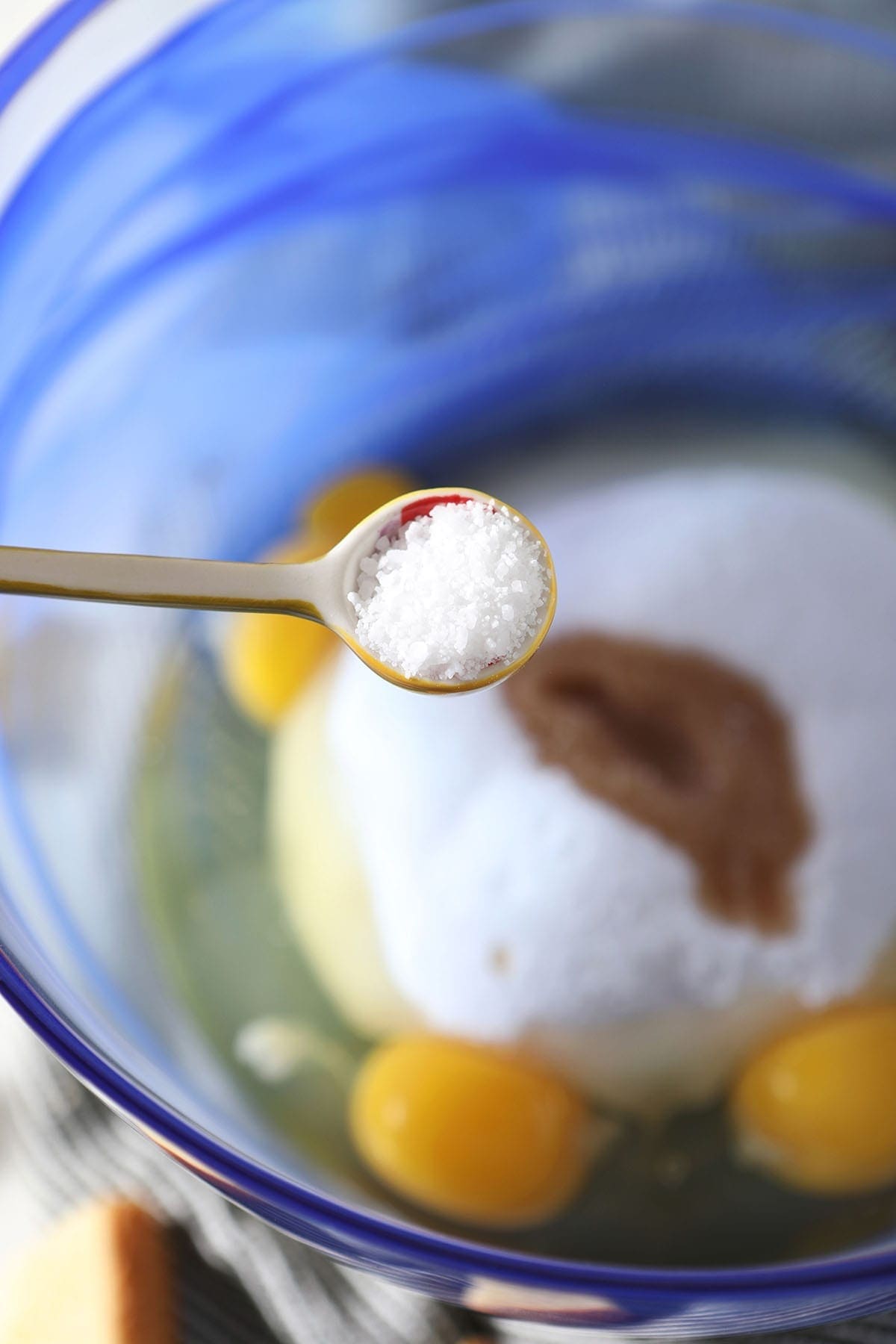 A teaspoon of salt sprinkles into a blue glass bowl holding eggs, sugar and vanilla extract