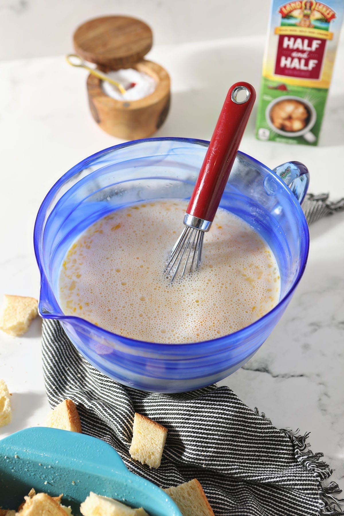 The custard is shown in a blue glass bowl with a whisk, surrounded by chopped bread, a canister of salt and a quart of Half & Half