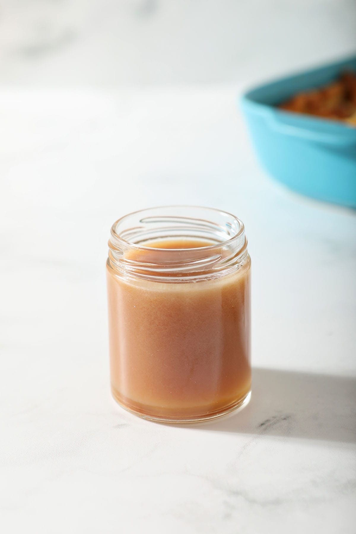 Caramel sauce in a jar sits on marble in front of a turquoise baking dish of Bread Pudding