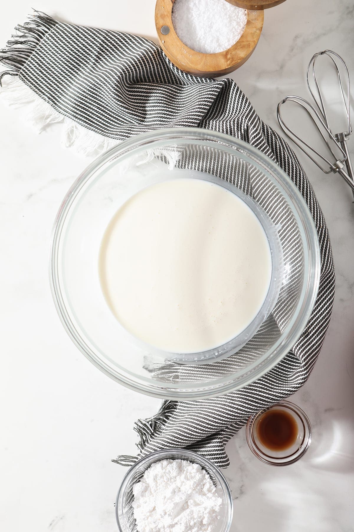 Overhead of a bowl holding heavy whipping cream sitting on a gray striped towel surrounded by powdered sugar, vanilla extract and salt