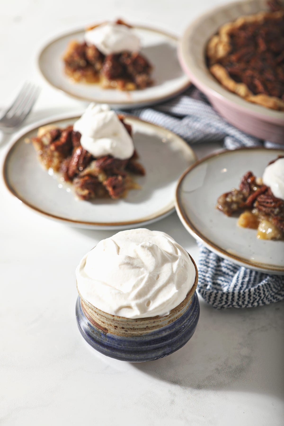 A blue pottery bowl holding a serving of homemade sweetened whipped cream next to three plates holding slices of pecan pie