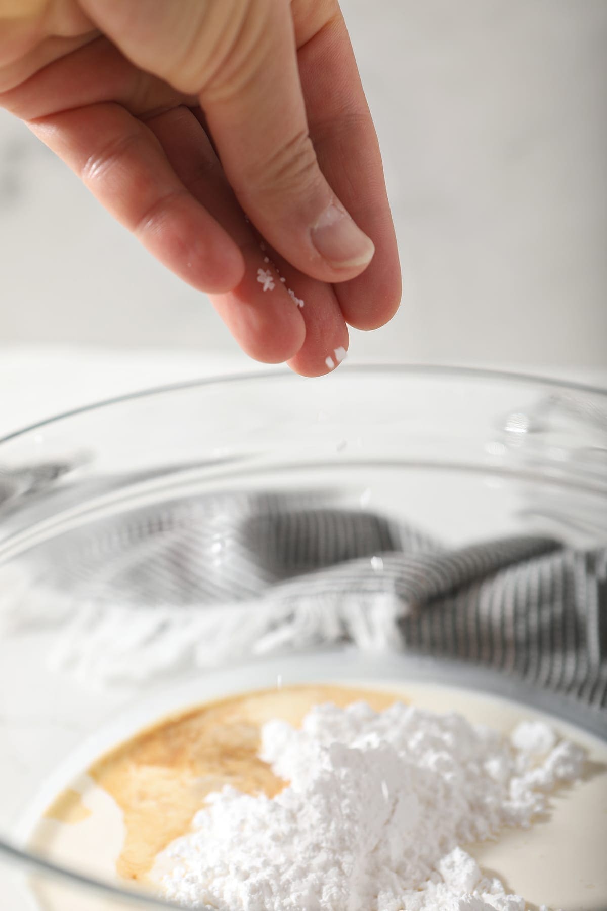 A hand sprinkles salt into a bowl holding cream, powdered sugar and vanilla