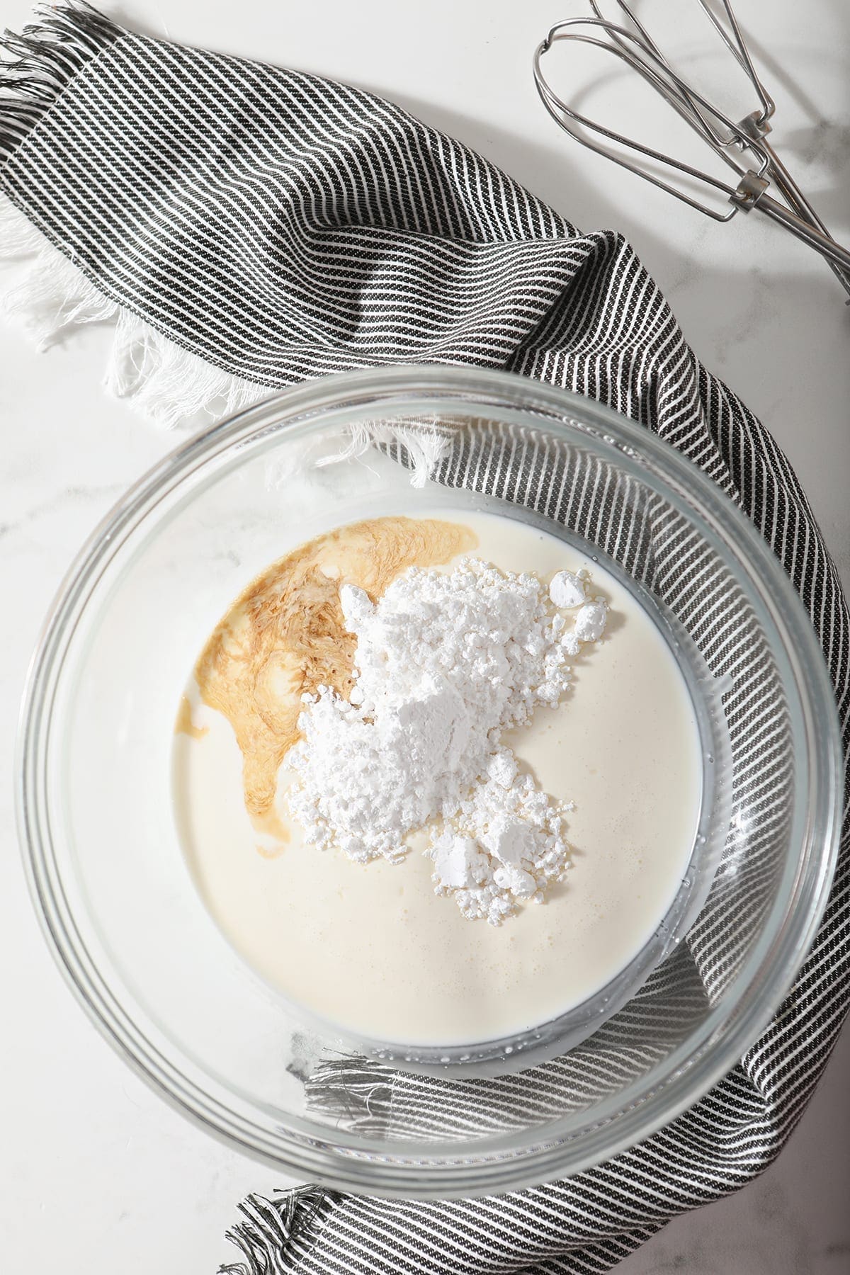 Homemade whipped cream ingredients in a bowl sit on a gray striped towel next to two hand mixer whisks