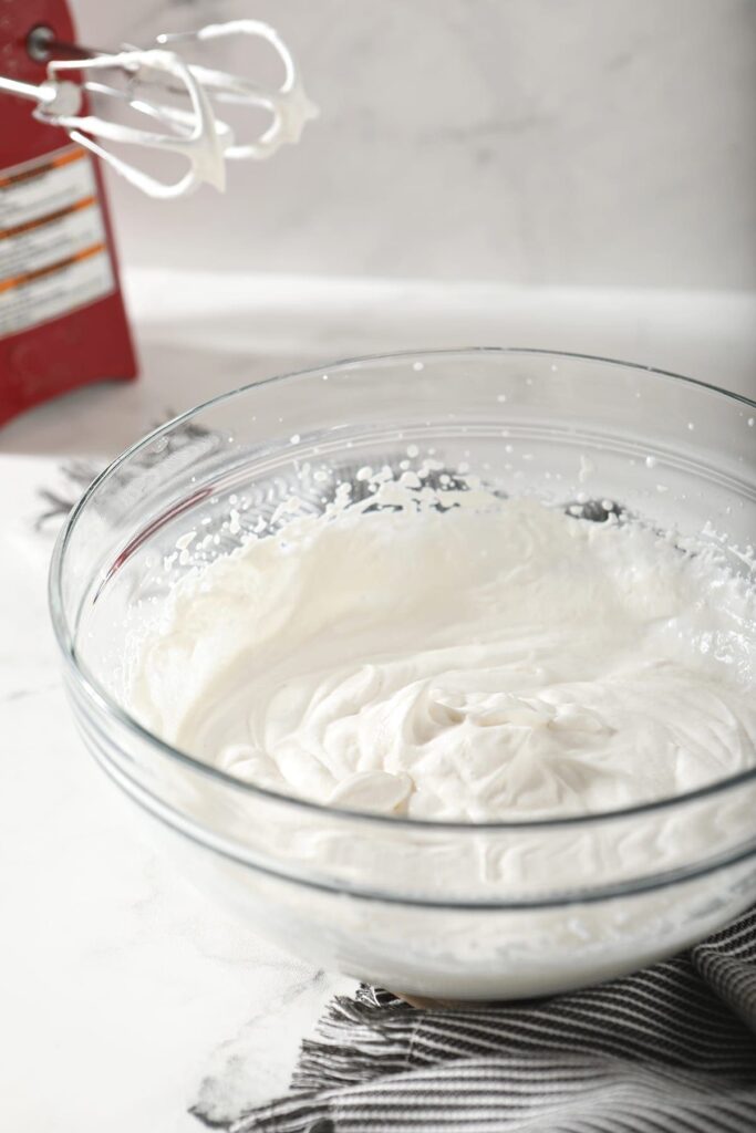 A bowl of homemade whipped cream sits in front of a red hand mixer