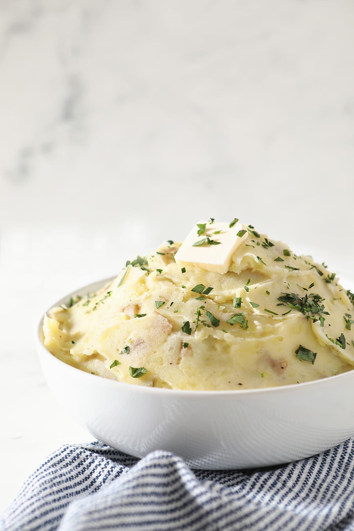 A white bowl holds a serving of creamy Homemade Mashed Potatoes while sitting on a blue and white striped towel