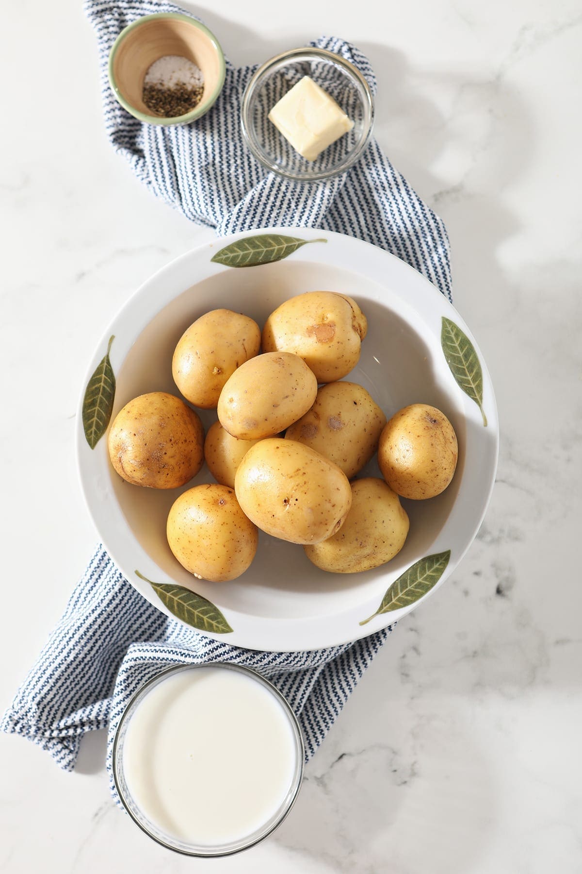 Gold potatoes sit in a decorative bowl on top of a blue and white striped towel next to a bowl of milk, butter and seasonings