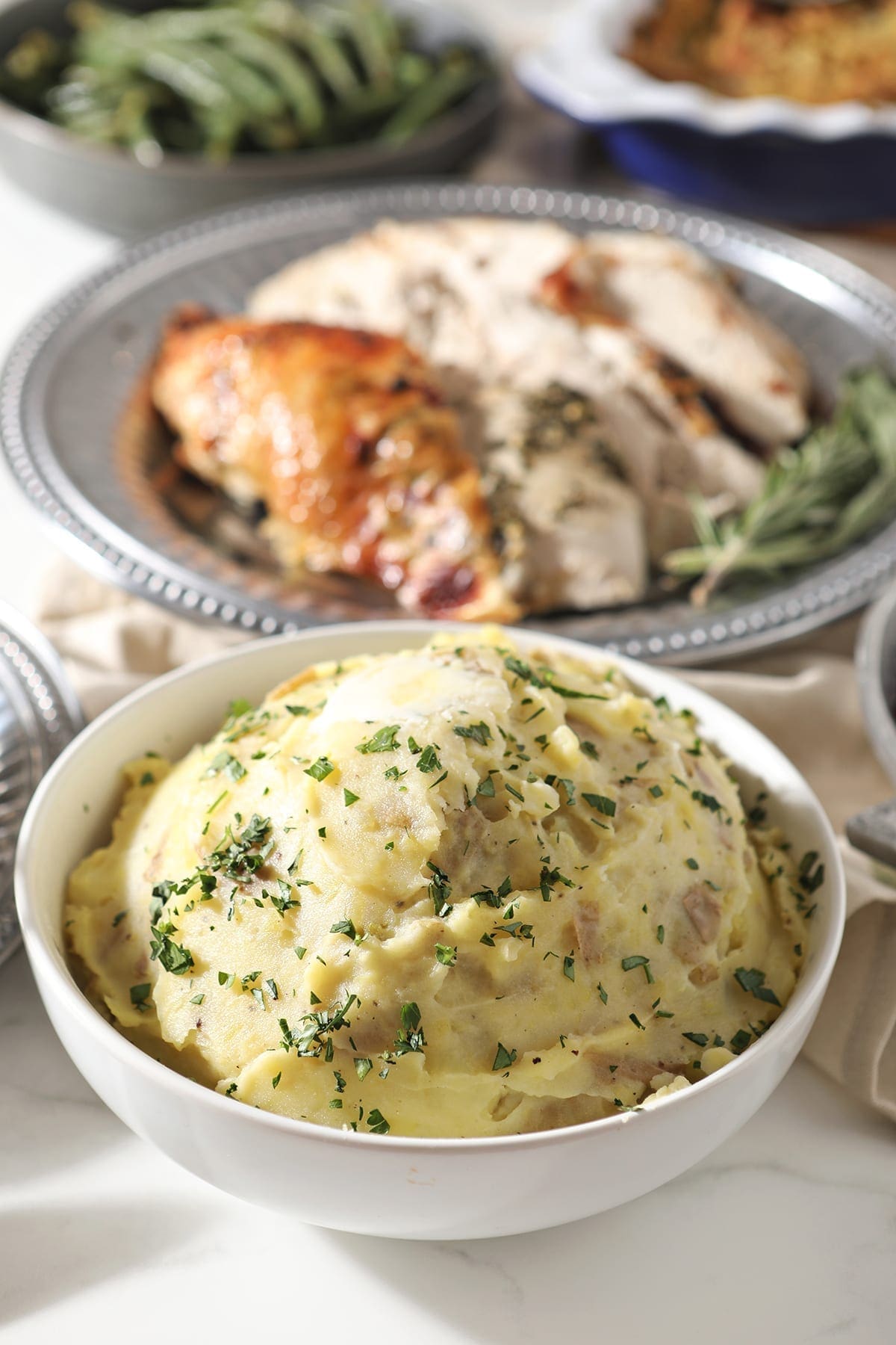 A bowl of mashed potatoes sits in front of a silver platter holding turkey, surrounded by other holiday dishes