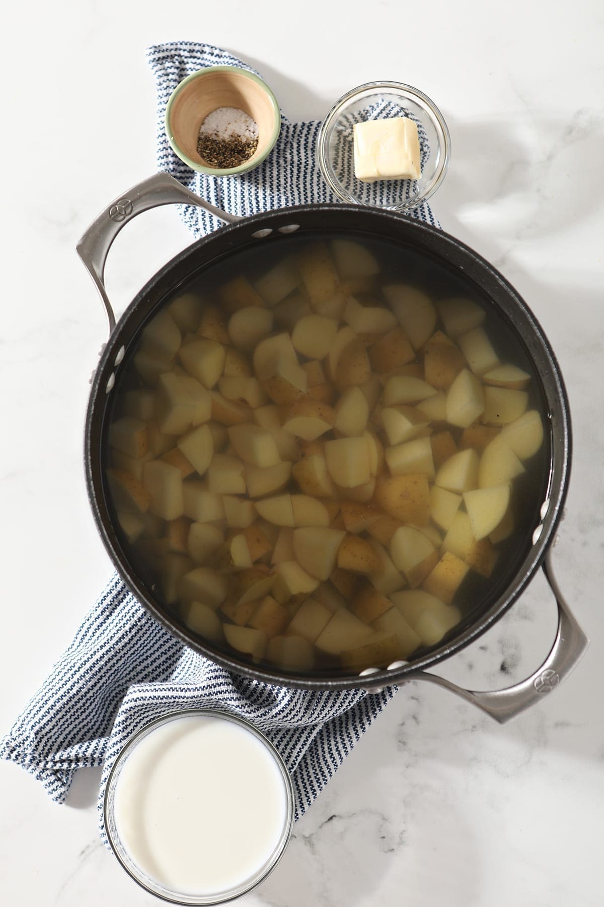 A large pot holding chopped potatoes sits next to other mashed potato ingredients while sitting on top of a blue and white striped towel