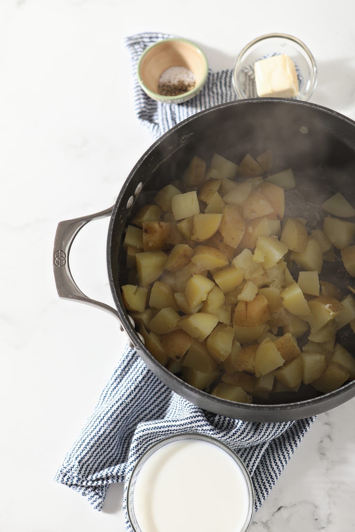 Boiled potatoes sit in a black pot next to bowls of milk, butter and seasonings