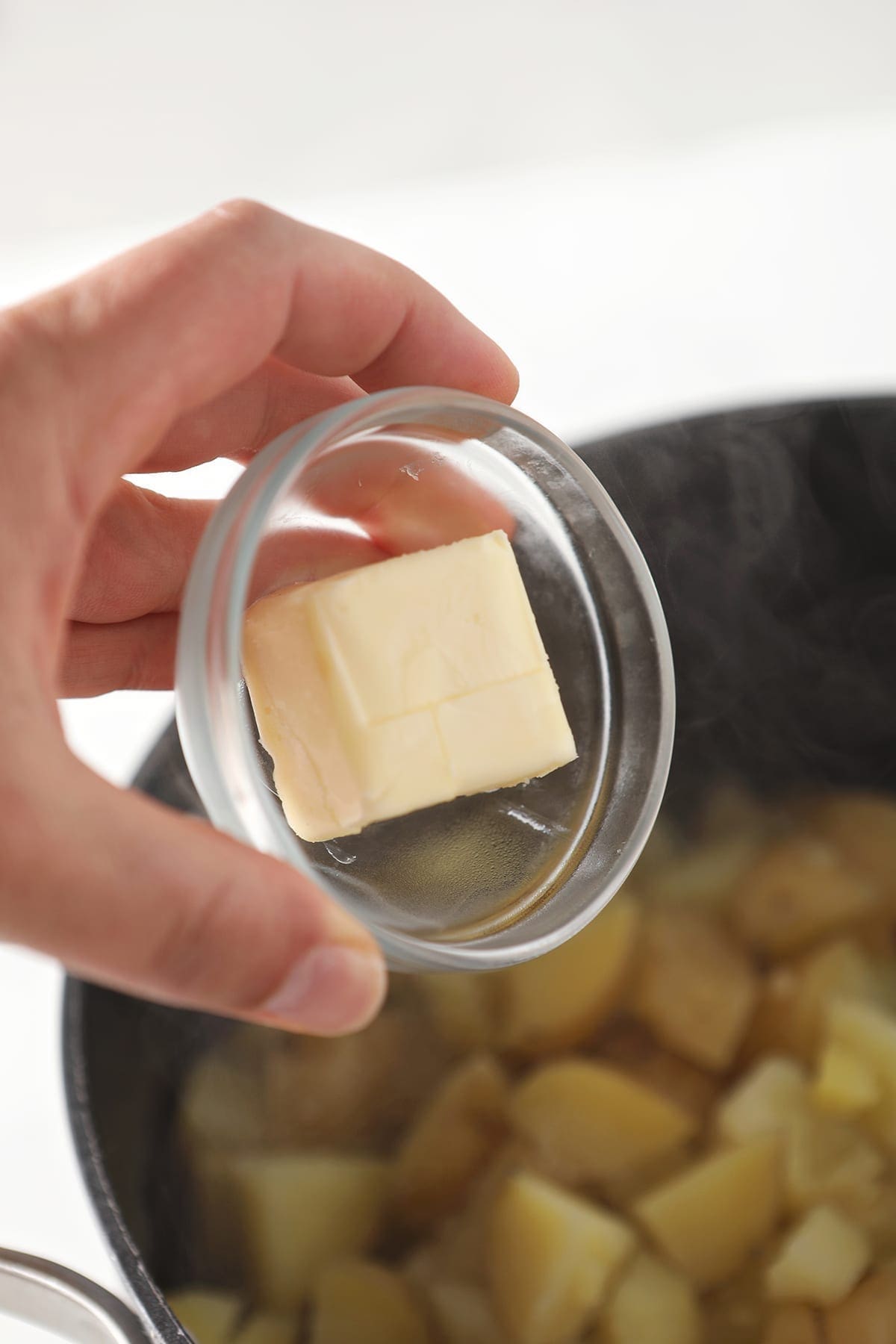 A bowl of softened butter is held over a pot holding boiled potatoes