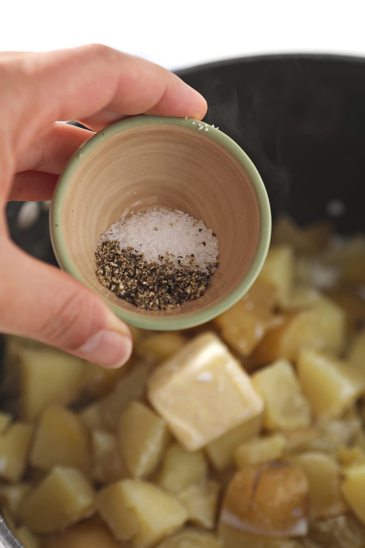 Salt and pepper are sprinkled from a bowl into a pot holding potatoes and other ingredients