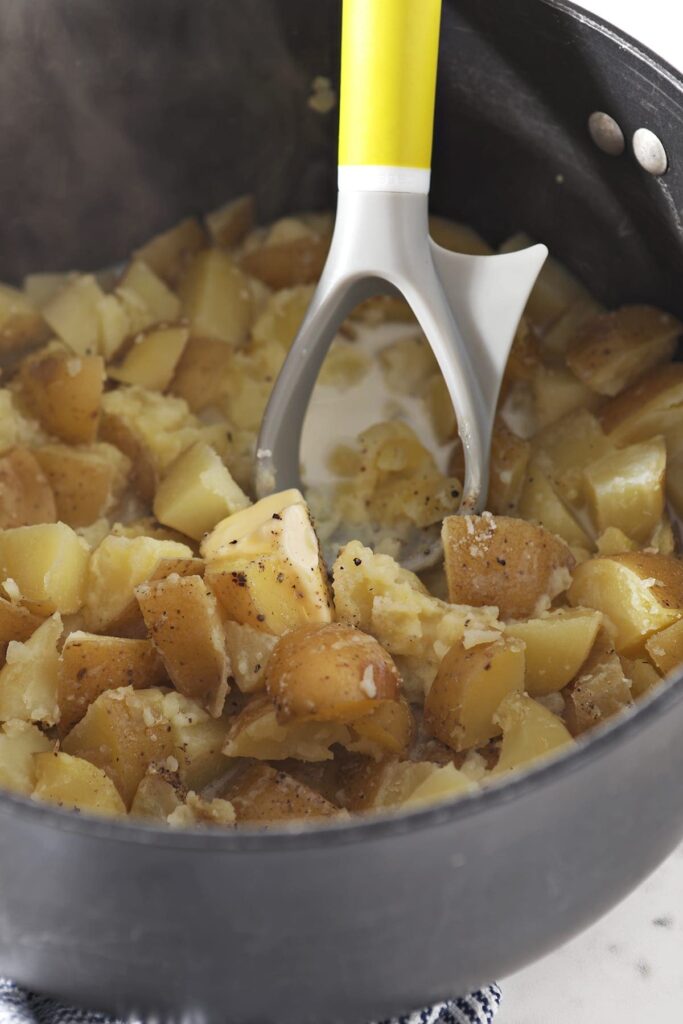 A potato masher is shown in a black pot with boiled potatoes and other ingredients