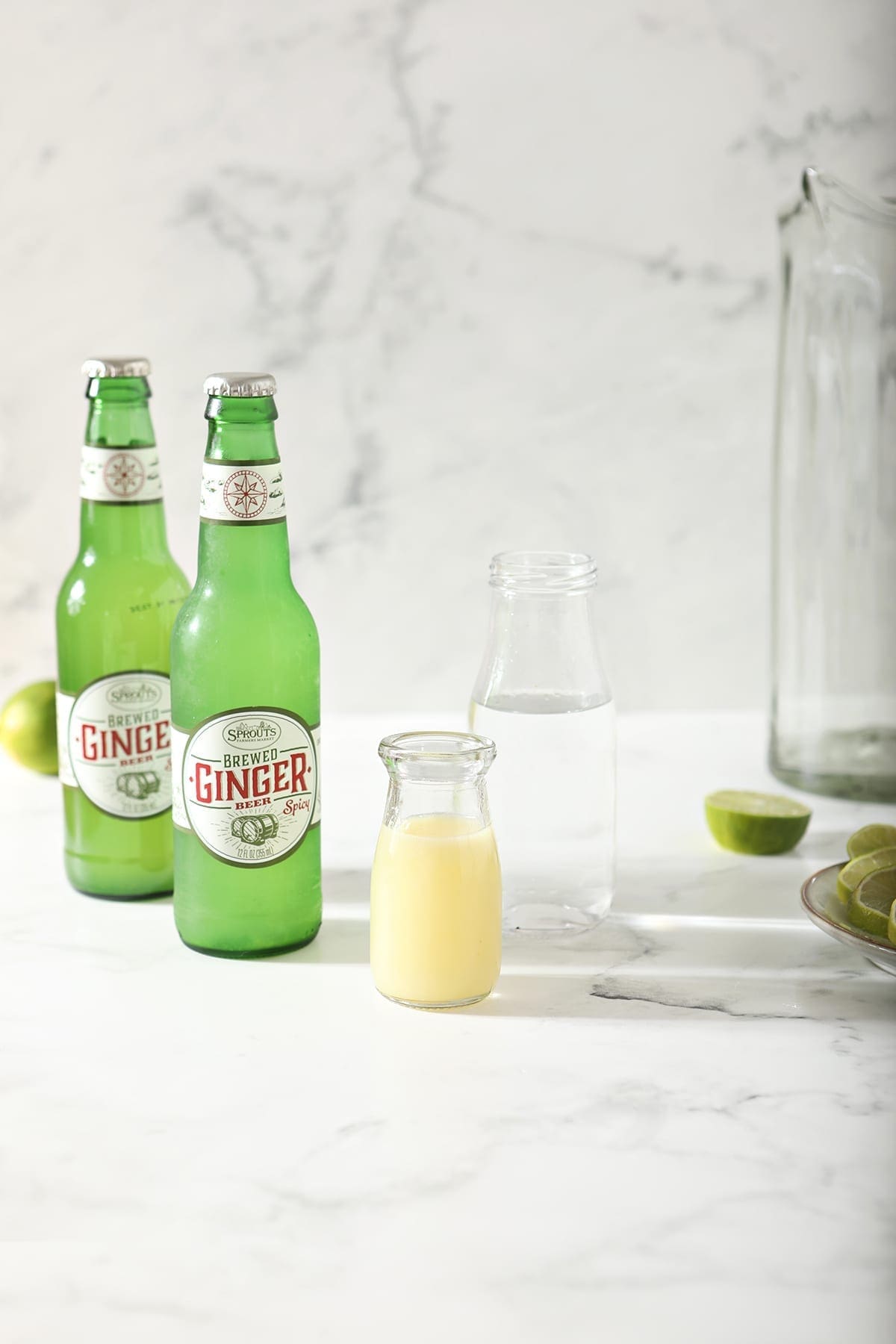 A bottle of lime juice with a bottle of vodka and two bottles of ginger beer on marble next to an empty pitcher and halved limes