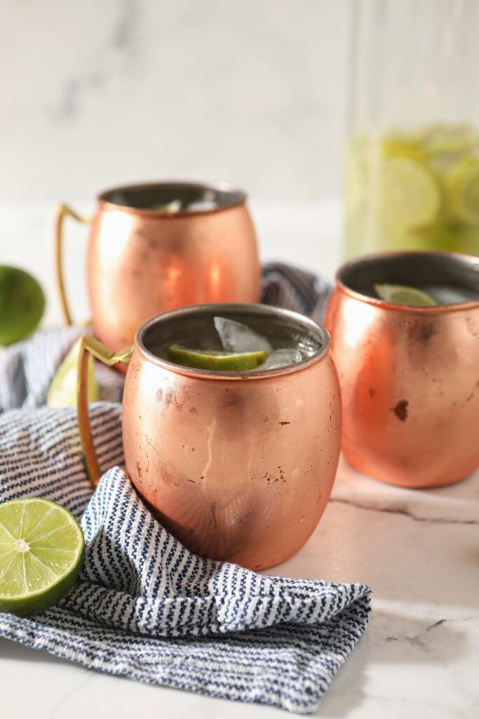 Three copper mugs sitting on a blue and white striped towel hold Moscow mules garnished with lime wedges on marble with a pitcher behind them